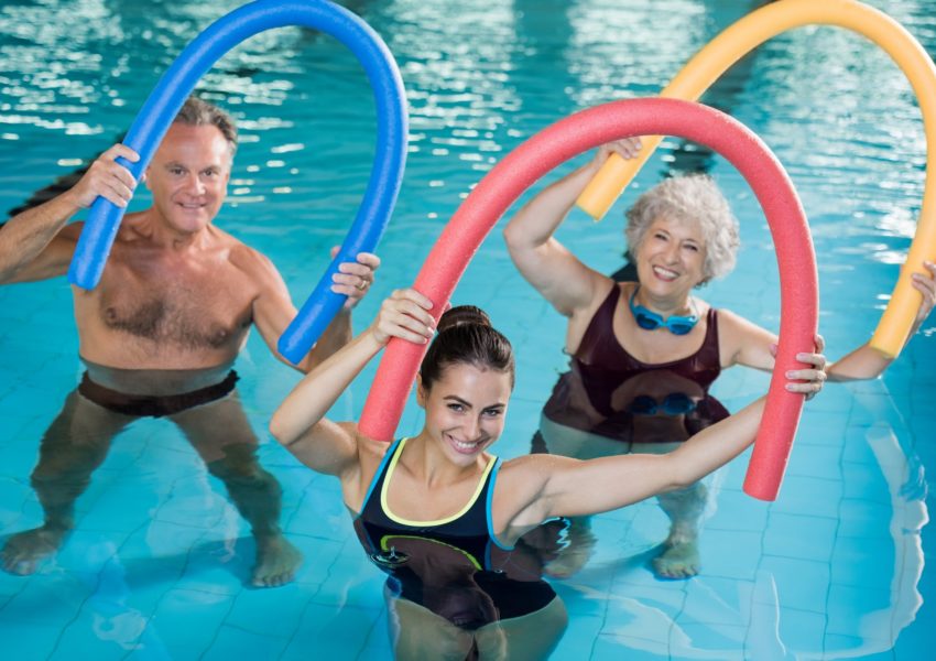 Portrait of smiling people doing aqua fitness together in a swimming pool. Group of senior woman and mature man with swim noodles exercising in a swimming pool. Young trainer and senior people in aqua gym fitness class.